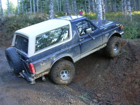 1991 Ford Bronco - Dark blue Ford Bronco with a white hardtop climbing a muddy dirt bank on a forest trail, viewed from the driver-side rear three-quarter wit…