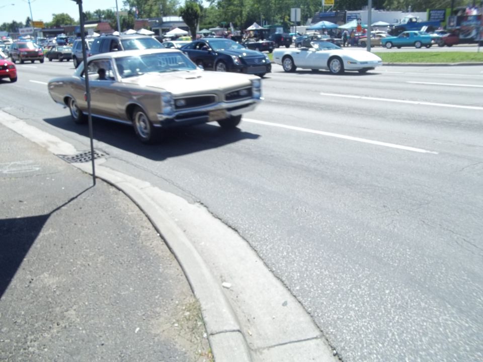 Gold 1966 Pontiac GTO driving on a sunny day, other cars and tents in background