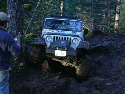 1991 Ford Bronco - A white Jeep Wrangler navigates a muddy trail, its front tires deep in the muck.