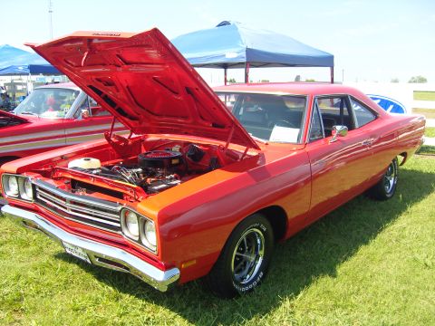 2007 Ford Mustang - A bright red 1969 Plymouth Road Runner is displayed with its hood open, revealing the engine bay. Another red car is visible behind it.