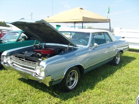 2007 Ford Mustang - A light blue 1965 Oldsmobile Cutlass is displayed with its hood open at an outdoor car show.