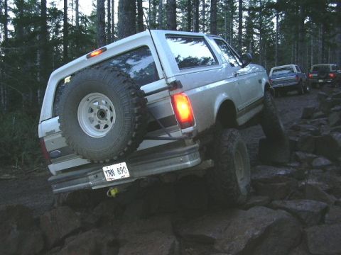 1991 Ford Bronco - White lifted Ford Bronco flexing over a rock ledge on a wooded trail, shot from the rear three-quarter with the passenger-side wheel droope…