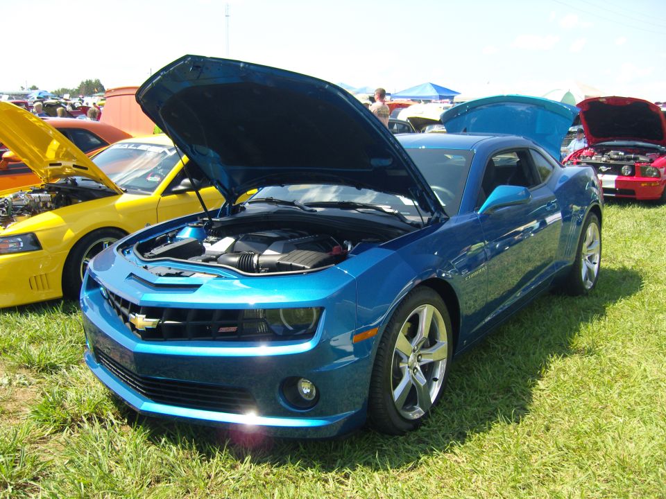 Blue Chevrolet Camaro parked on grass with hood open, yellow Ford Mustang and red car in background