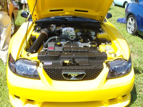 2007 Ford Mustang - The engine bay of a yellow 2007 Ford Mustang is displayed with the hood open, revealing a supercharger and other engine components.