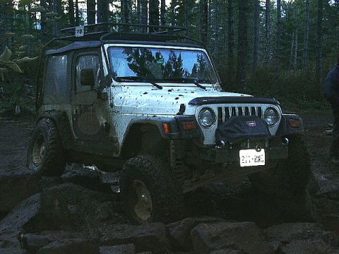 1991 Ford Bronco - A white Jeep Wrangler navigates a rocky trail in a wooded area, with mud splattered on its body.