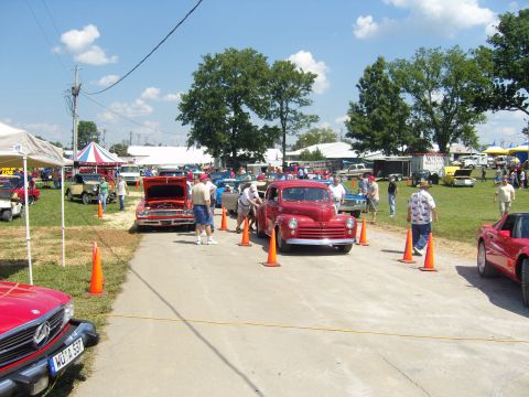 2007 Ford Mustang - A red vintage car is parked at a car show, with other vehicles and tents visible in the background.