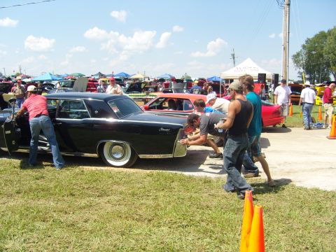 2007 Ford Mustang - Several people are pushing a large black classic car at an outdoor event with many other vehicles and tents visible.
