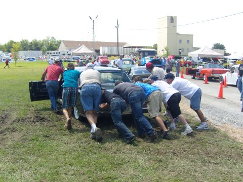 2007 Ford Mustang - Several people are pushing a car at an outdoor event with other vehicles and tents in the background.