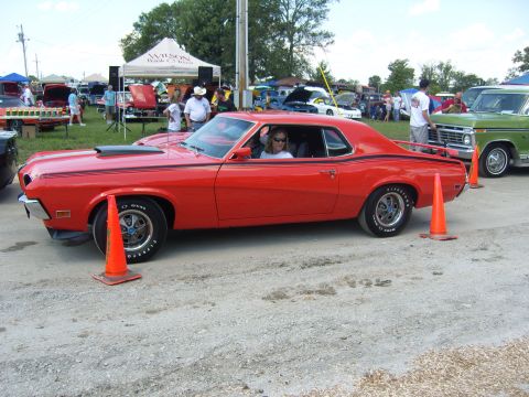 2007 Ford Mustang - A red coupe with black racing stripes is parked at an outdoor event, with other vehicles and tents in the background.