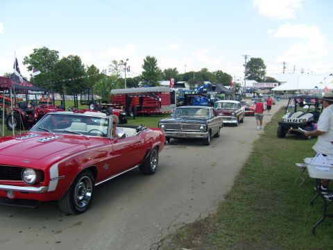 2007 Ford Mustang - A line of classic cars, including a red 1969 Camaro convertible, proceeds down a road at an outdoor event.
