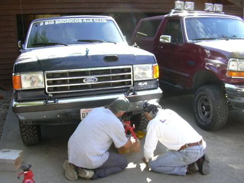 1991 Ford Bronco - Two people kneel on a driveway working under the front bumper of a dark green Ford Bronco with a "2 BIG BRONCOS 4x4 CLUB" windshield banner…