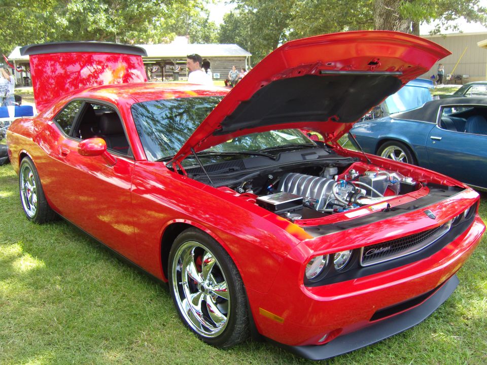 Red Dodge Challenger with hood and trunk open, showing engine and spoiler. Another blue car is parked behind it
