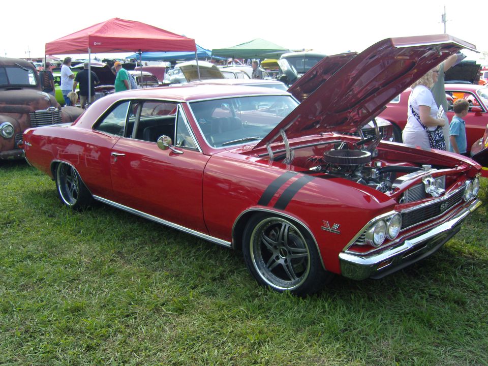Red classic car with black racing stripes on the hood and fender, hood open showing engine, parked on grass at an outdoor event