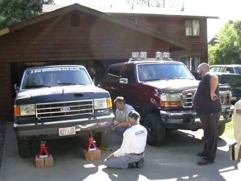1991 Ford Bronco - Two full-size Ford Broncos in a driveway: a black one on jack stands with a '2 Big Broncos 4x4 Club' windshield banner, beside a maroon lif…