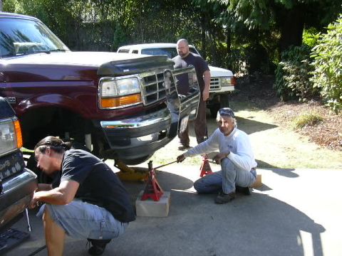1991 Ford Bronco - A maroon Ford Bronco sits nose-up on red jack stands and a floor jack in a driveway with its front wheel removed, while three men work arou…