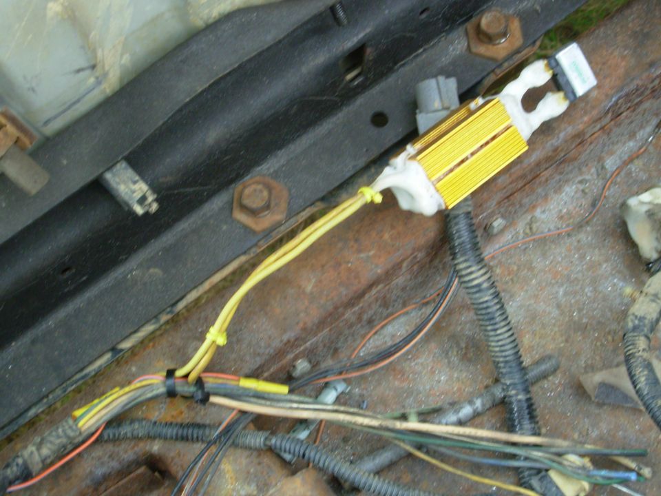 Gold finned load resistor wired with yellow leads on rusty vehicle surface with harnesses