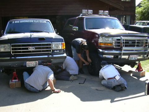 1991 Ford Bronco - Two 1990s full-size Ford Broncos parked nose-out in a driveway while four people work underneath them on jack stands, one dark blue with a…