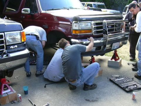 1991 Ford Bronco - Two people work on the front end of a maroon Ford Bronco or F-Series truck raised on red jack stands over wood blocks in a driveway. A sock…