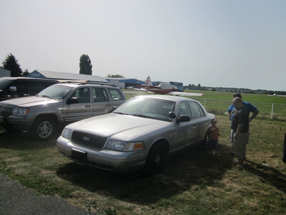 Silver sedan parked on grass, front quarter view. A Jeep Grand Cherokee is to its left. People stand near the sedan