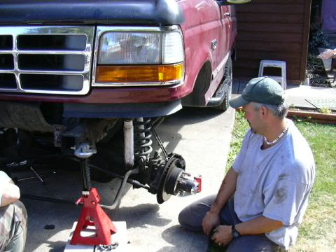 1991 Ford Bronco - Front suspension work in progress on a red Ford truck raised on a red jack stand in a driveway, with the wheel removed to expose the coil s…