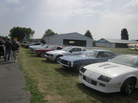 1991 Ford LTD Crown Victoria - A lineup of classic cars, including a white Camaro, parked outside hangars on a sunny day.