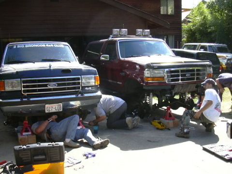 1991 Ford Bronco - Driveway wrenching session with a maroon full-size Bronco on jack stands, front wheels off and coil-spring solid front axle exposed, roof-r…