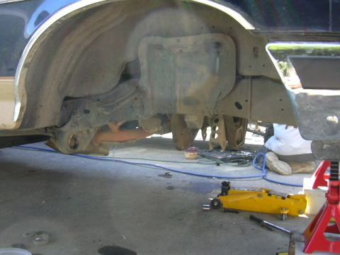 1991 Ford Bronco - Undercarriage and wheel-well of a blue vehicle up on stands during work, with the inner fender, frame rail and suspension exposed. A yellow…