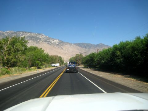 1996 Ford Bronco - A blue pickup truck tows a trailer down a highway, with mountains and trees lining the road under a clear blue sky.