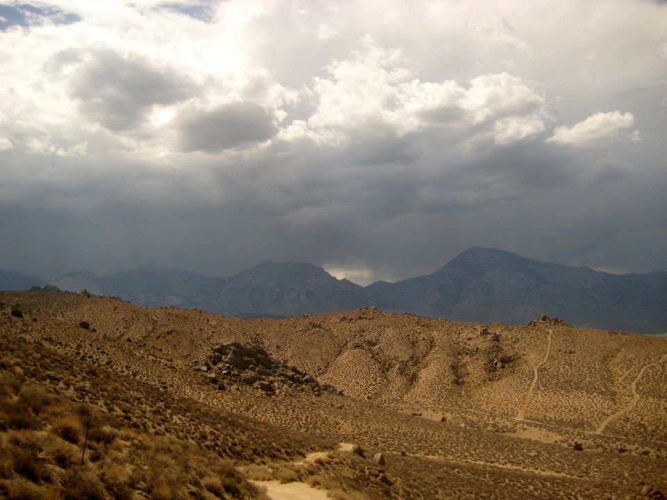 Desert landscape with dry brush, rocky hills, and distant mountains under a cloudy sky
