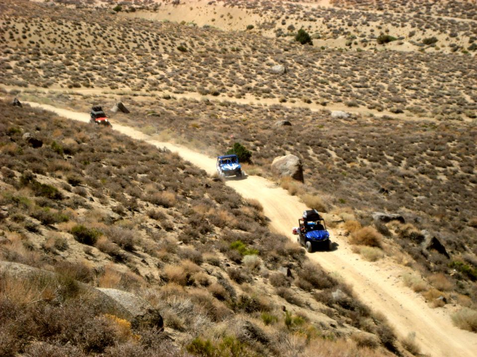 Three off-road vehicles travel on a dusty dirt road through a dry, brush-covered hillside