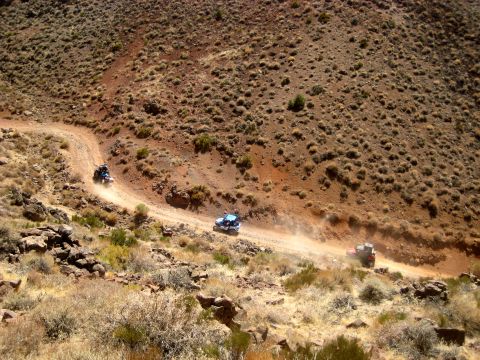 1996 Ford Bronco - Multiple ATVs traverse a dusty, winding dirt road through a barren, arid landscape.