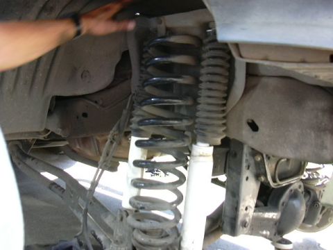 1991 Ford Bronco - Close-up under a truck's front fender showing a black lift coil spring and a white-bodied shock absorber with a black boot, mounted alongsi…