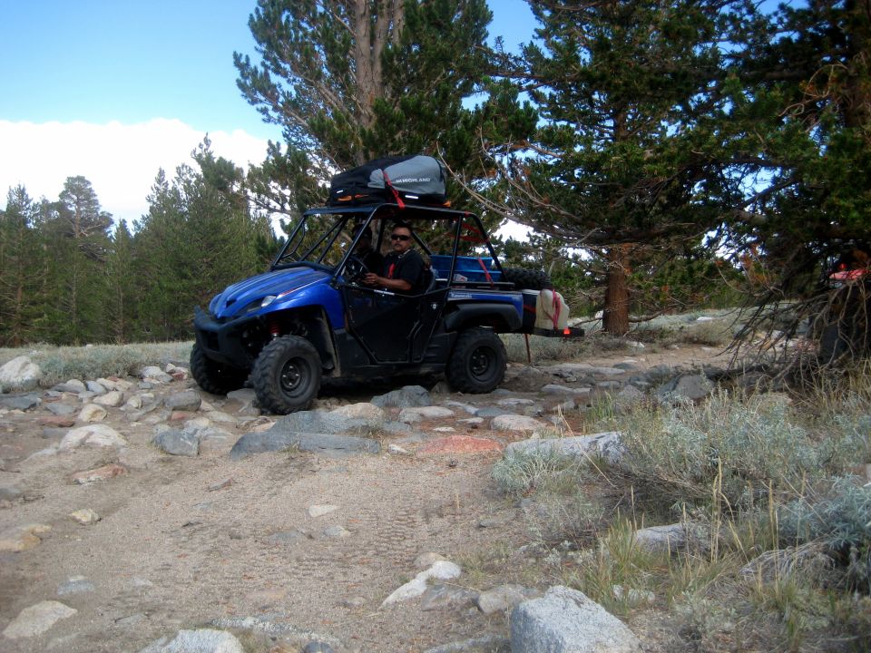 Blue side-by-side ATV with luggage on roof rack driving on a rocky mountain trail