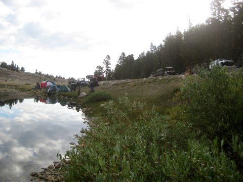 1996 Ford Bronco - A group of off-road vehicles and tents are camped by a lake, with the sky reflected in the calm water.