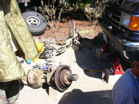 1991 Ford Bronco - Driveway suspension teardown with a removed front axle assembly, brake rotor and hub laid on the concrete alongside coil springs, radius ar…