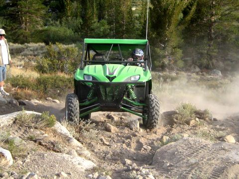 1996 Ford Bronco - A person drives a green off-road vehicle through a dusty, rocky terrain with trees in the background.