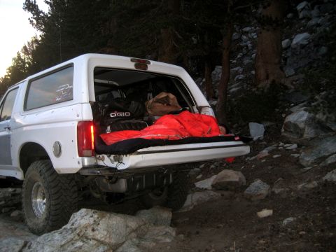 1996 Ford Bronco - A white Ford Bronco is parked on a rocky trail with its tailgate open, revealing camping gear and a sleeping bag.