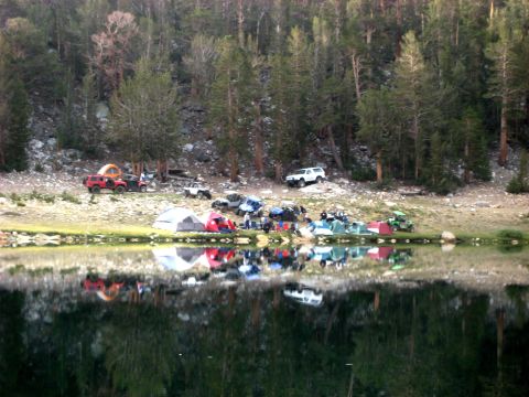1996 Ford Bronco - A group of off-road vehicles and tents are set up near a reflective lake in a wooded area.