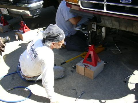 1991 Ford Bronco - Two men work on the front end of a dark-red Ford truck raised on a red jack stand set on a wood block, with a yellow floor jack, breaker ba…
