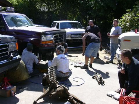 1991 Ford Bronco - Driveway wrenching session with a dark maroon full-size Ford truck up on jack stands, front wheel removed and several people working on and…