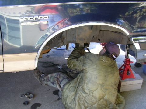 1991 Ford Bronco - A person in camo pants lies on a padded moving blanket working under the empty rear wheel well of a dark blue Ford Bronco, with the BRONCO…