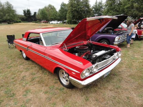 1964 Ford Fairlane - A red classic coupe is displayed with its hood open at an outdoor event, alongside other vehicles.