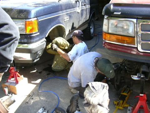 1991 Ford Bronco - Two people work between a blue and a maroon 1980s-90s Ford truck/SUV parked nose-out in a driveway, one crouched at the blue truck's open f…