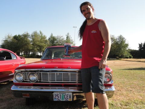 1964 Ford Fairlane - A man poses with a trophy in front of a classic red car, with another vehicle partially visible.