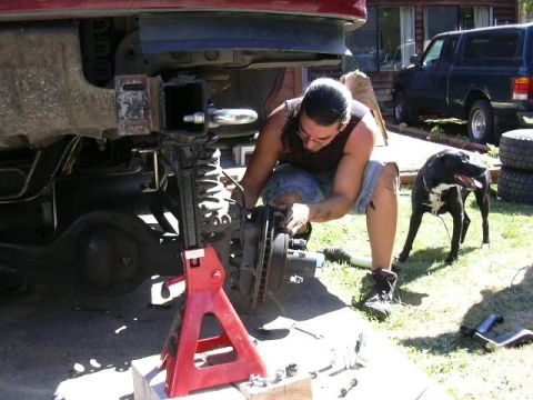 1991 Ford Bronco - A person in a sleeveless shirt kneels in a sunlit yard working on the front brake and hub of a red truck raised on a red jack stand, with t…