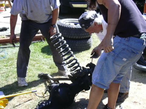 1991 Ford Bronco - Two people work on a black-painted solid front axle lying on the grass in a yard, with a tall coil spring standing on the axle's spring per…