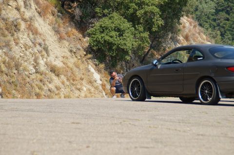 1993 Ford Taurus - A dark-colored coupe with aftermarket black rims is seen from the side on a paved road, with a man crouching in the background.