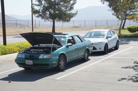 1993 Ford Taurus - Two cars parked in a lot: a green 1993 Ford Taurus with its hood up and a white hatchback.