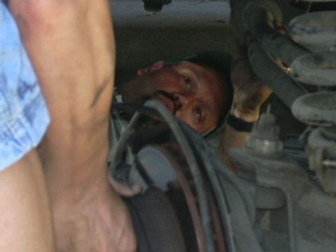 1991 Ford Bronco - A person lying under a raised truck looks up from beside the front suspension, with a coil spring, brake drum or rotor and brake hose visib…
