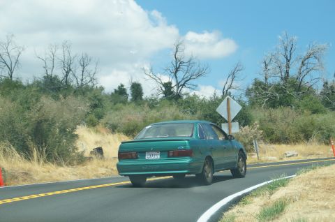 1993 Ford Taurus - Rear view of a green 1993 Ford Taurus driving on a highway with a scenic, dry landscape.